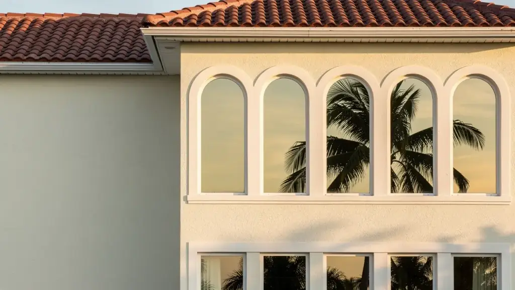 Arched second-floor windows on a cream stucco Naples house, the clean glass holding a palm reflection at golden hour