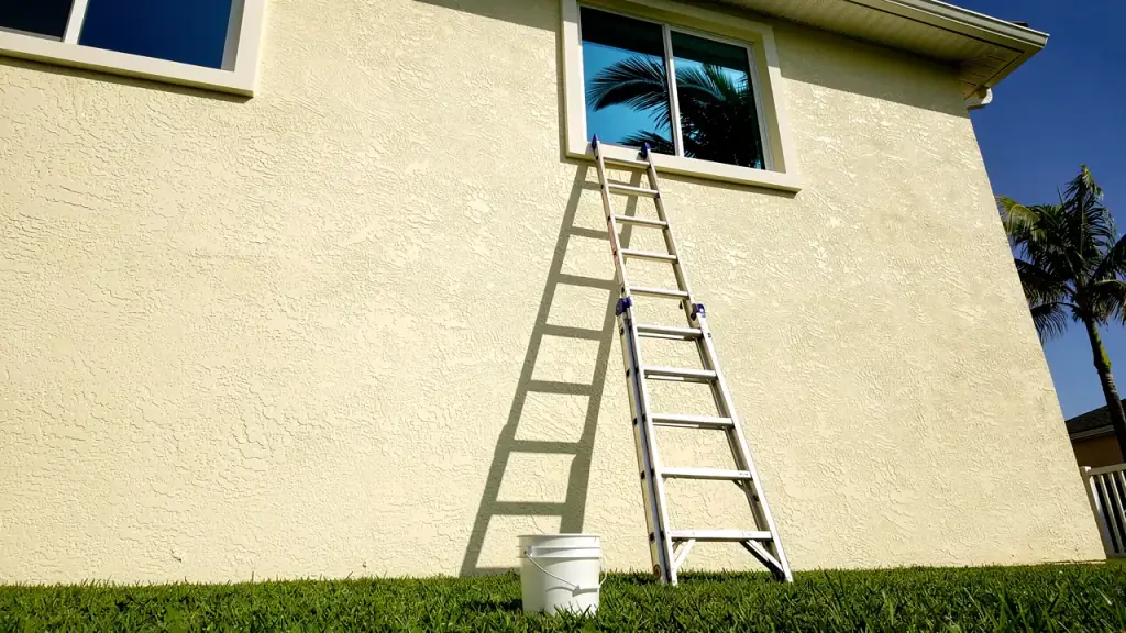 An aluminium ladder against a cream stucco wall, its top just under a clean second-floor window reflecting a palm and blue sky